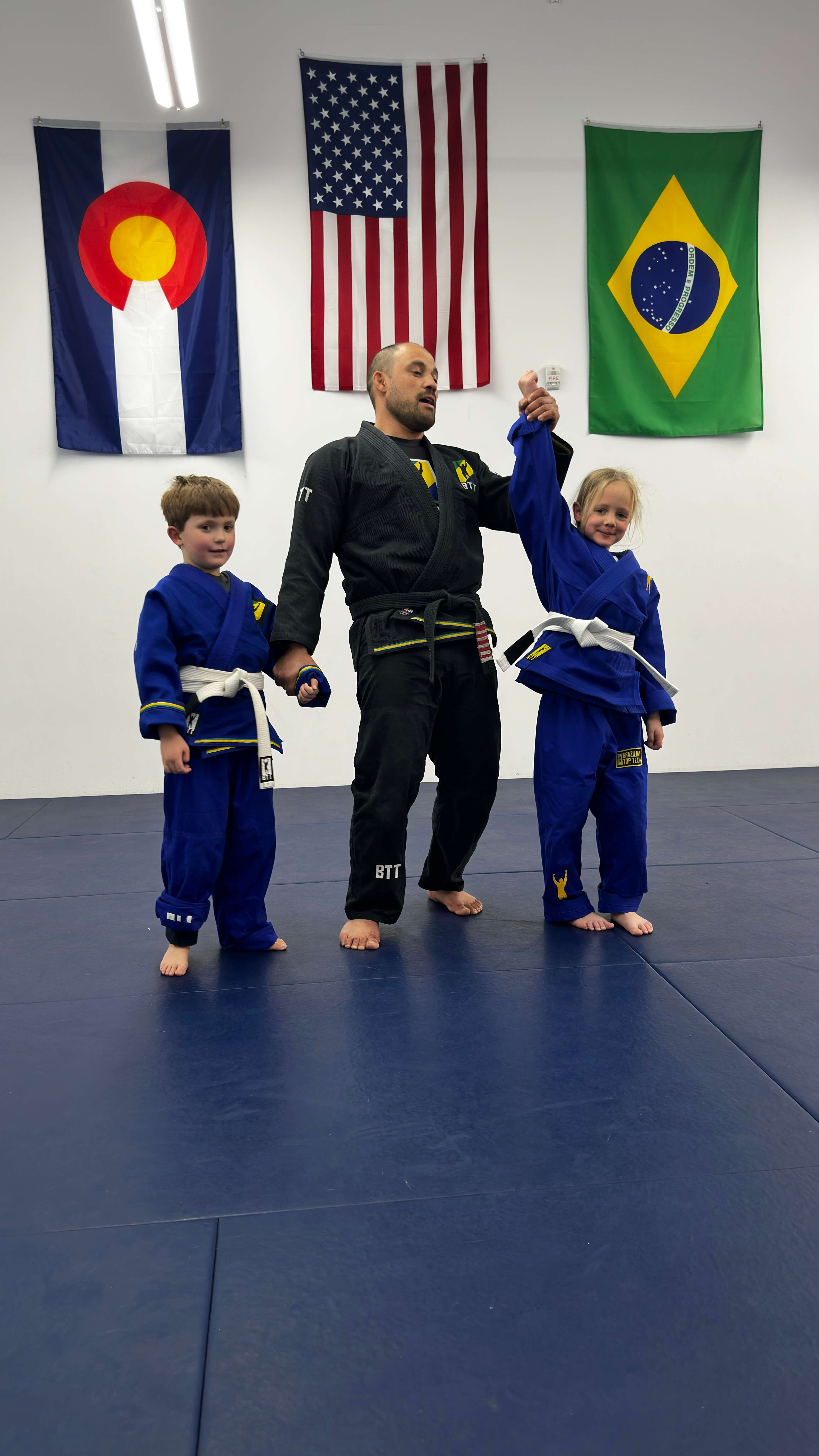 Martial arts instructor raises a girl's arm in victory beside a boy on blue mats.