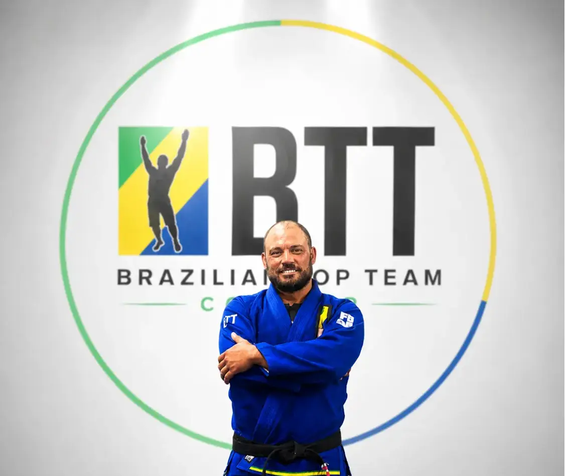 Smiling martial artist in blue gi and black belt posing before Brazilian Top Team logo.